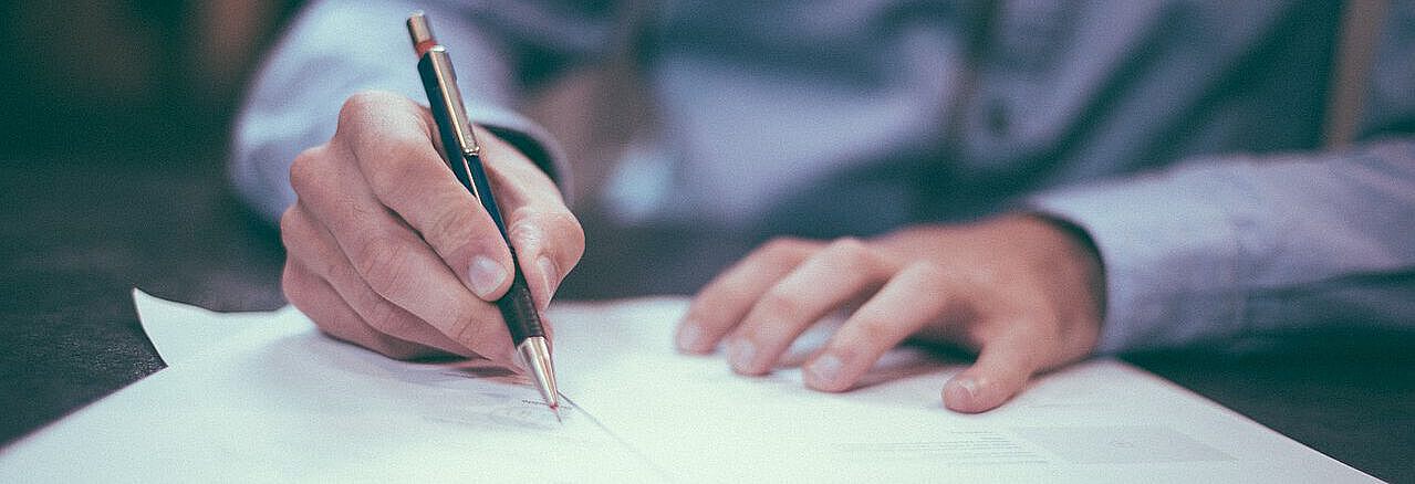 Symbolbild einer Person die gerade arbeitet: Eine Person hat einen Stift in der Hand, vor ihr liegen mehrere Blätter Papier, auf welche die Person mit ihrem Stift zeigt