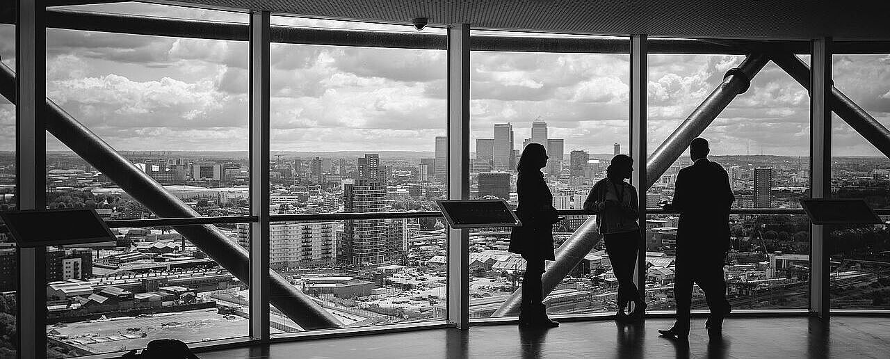 Schwarz-Weiß-Bild: Drei Personen unterhalten sich in einem Hochhaus vor einer Glasfassade, im Hintergrund ist die Skyline einer Großstadt zu sehen
