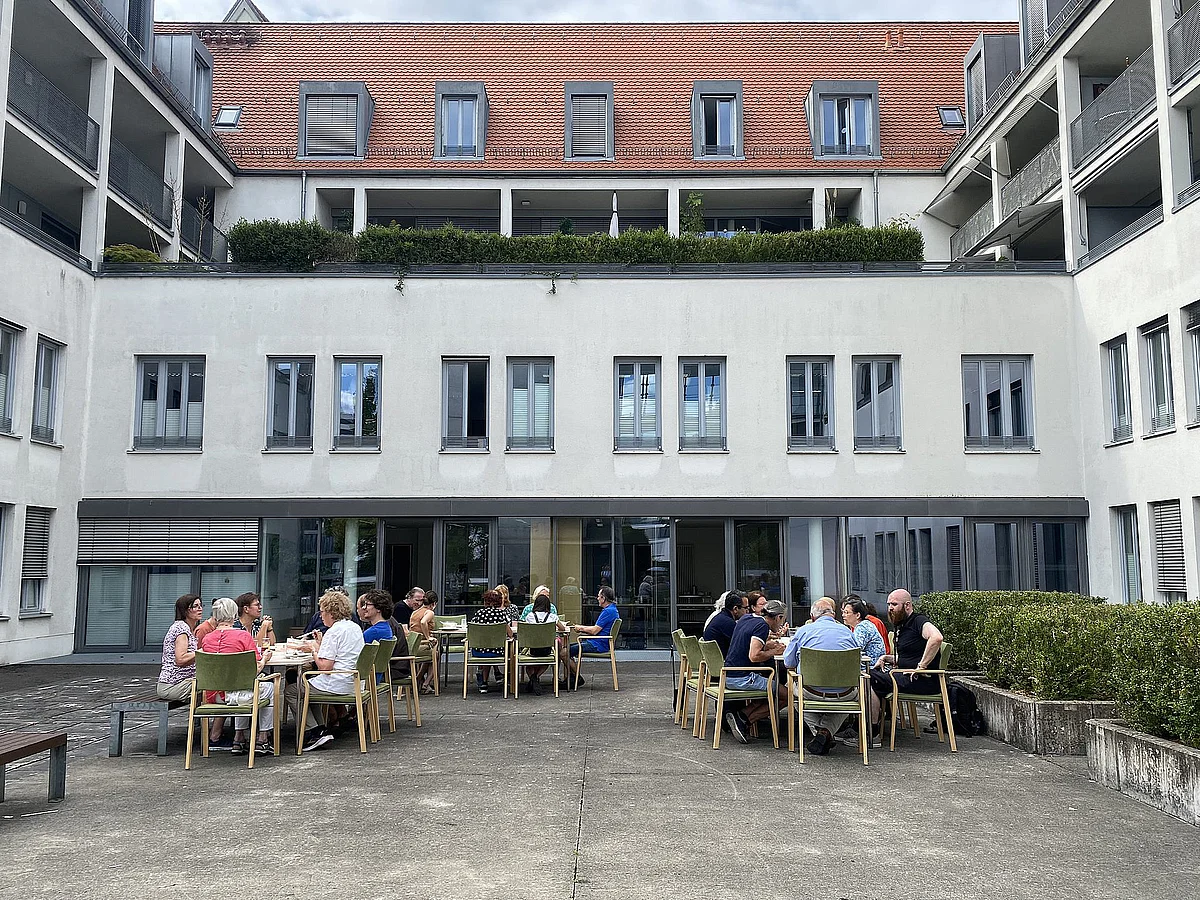 An drei Tischen sitzen Teilnehmende miteinander im Innenhof, essen und reden gemeinsam.