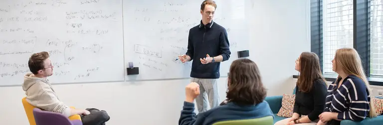 Studierende sitzen in Sesseln und hören einem Dozenten zu, der an einem Whiteboard steht