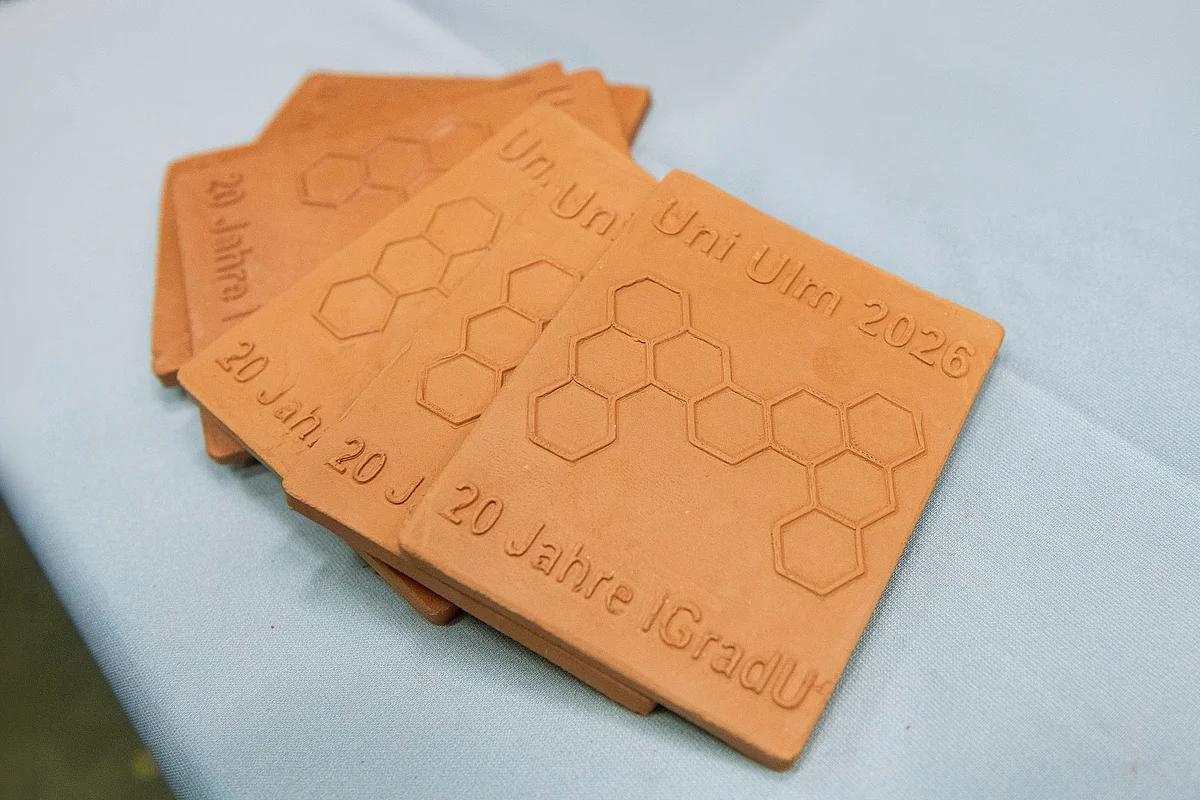 Several small ink tablets are arranged in a fan-like pattern next to one another. They show a molecular structure and the text ‘University of Ulm 2026 – 20 years of IGradU’