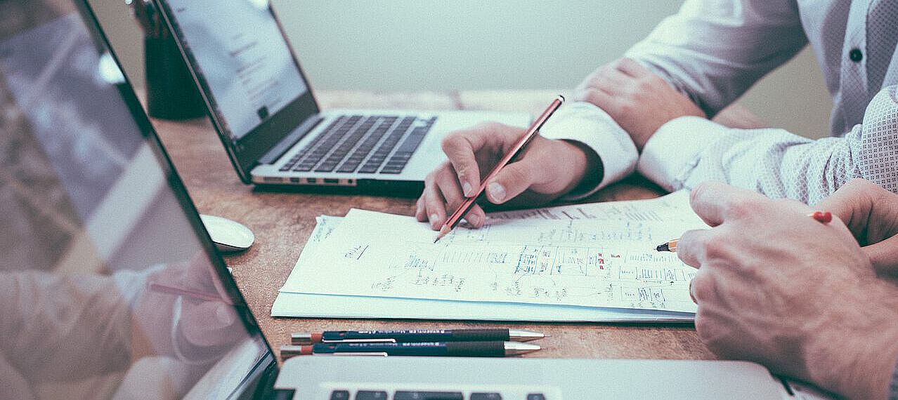 Symbolbild zweier arbeitenden Personen: Zwei Personen sitzen mit ihren Laptops vor einigen Blättern Papier, Person 1 zeigt Person 2 etwas darauf