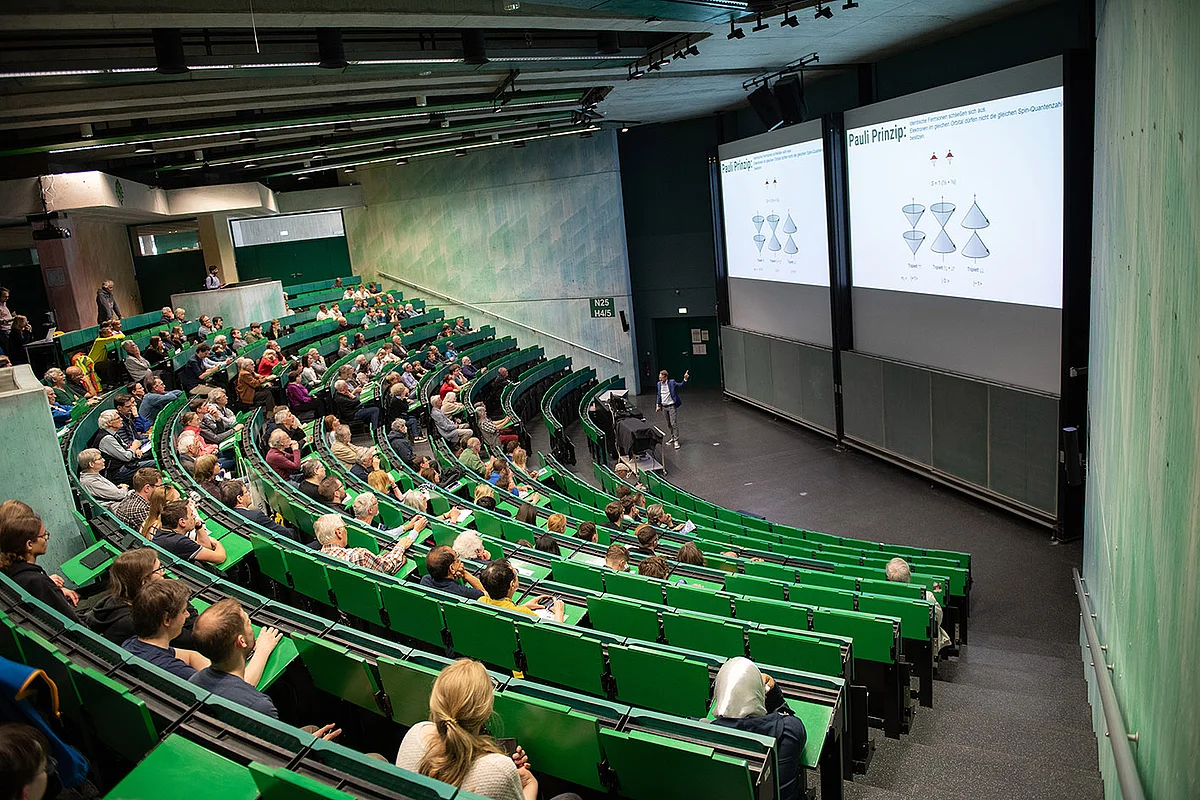 In gut gefüllter Hörsaal mit grünen Banken, ein Wissenschaftler präsentiert Folien