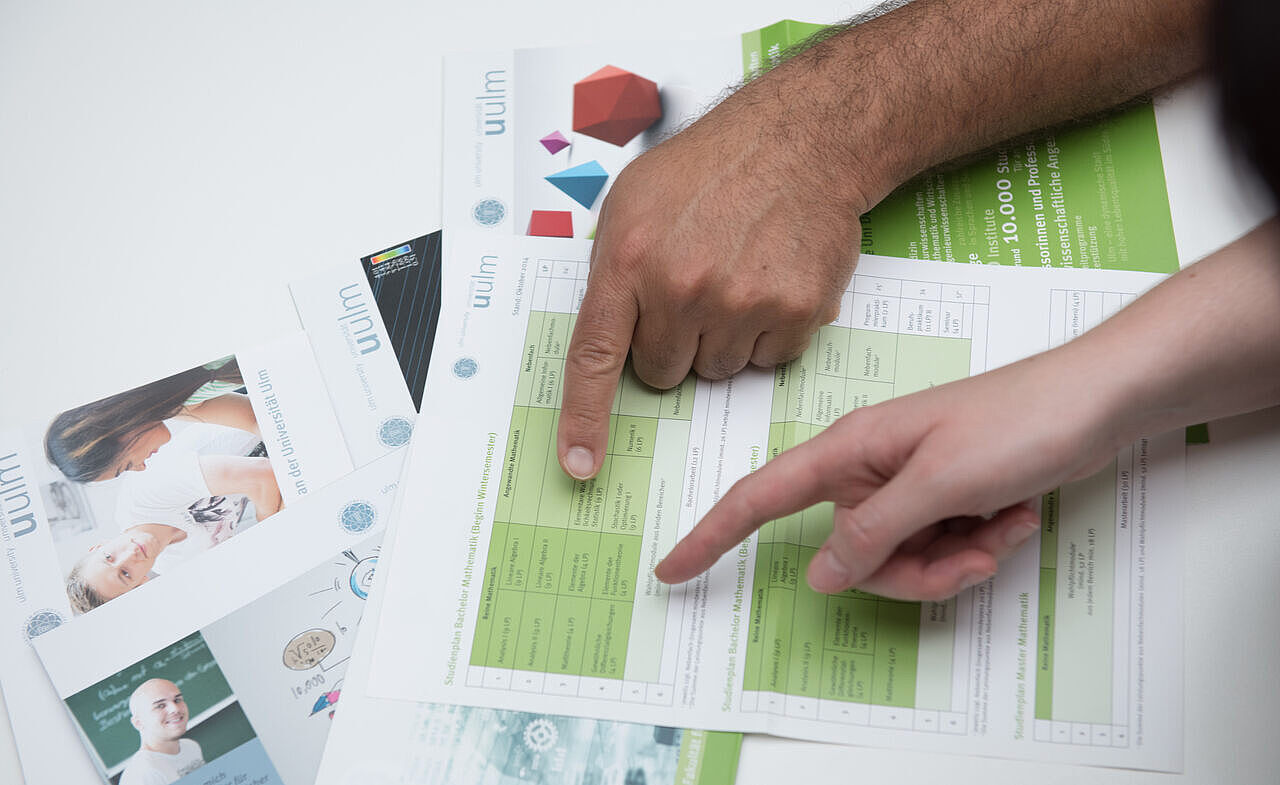 Während der Studienberatung zeigt ein Berater auf einen Studienplan. Im Bildausschnitt sind zwei Hände und verschiedene Flyer zu sehen. 
