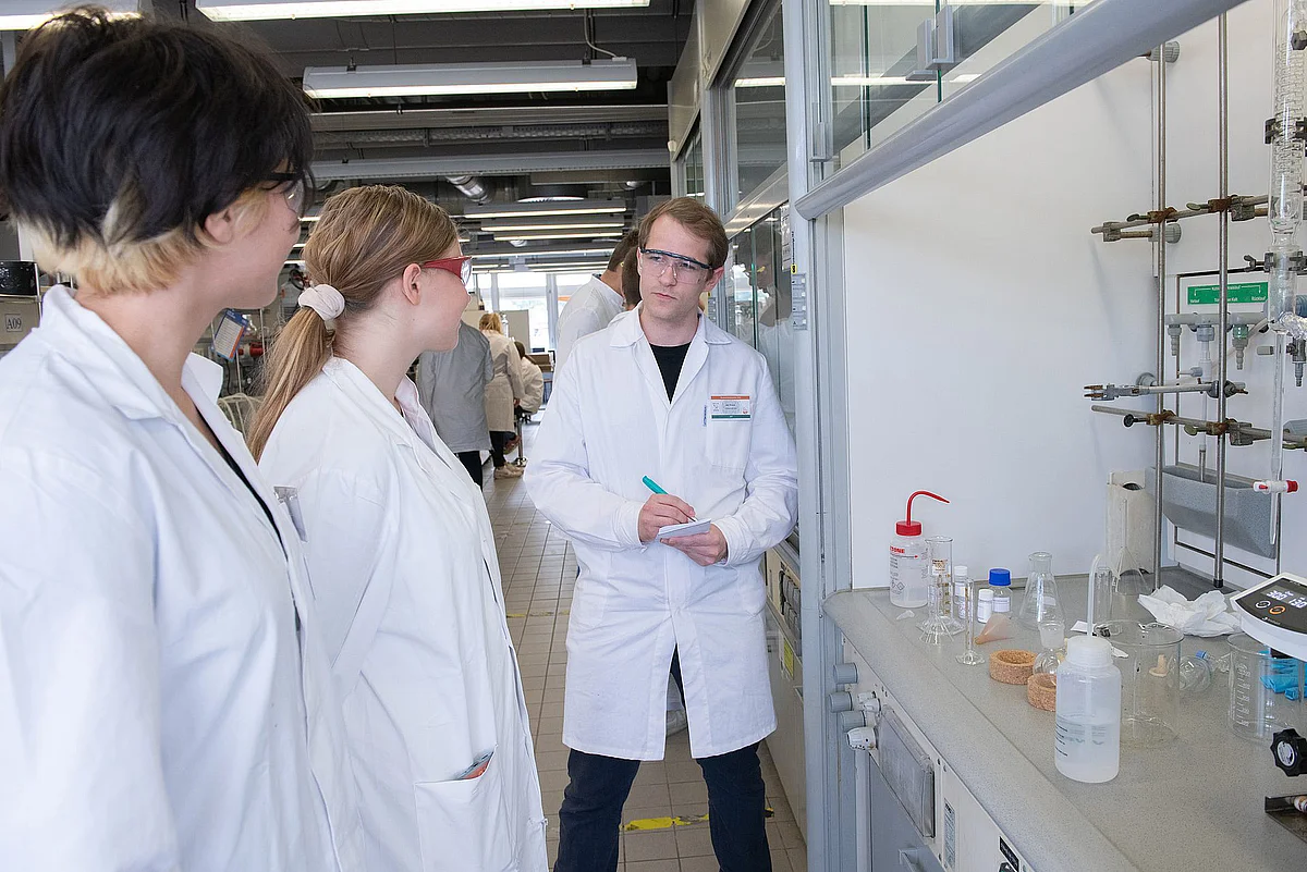 In einem Chemielabor hören zwei junge Frauen hören aufmerksam einem Mann zu. Alle drei tragen weiße Laborkittel