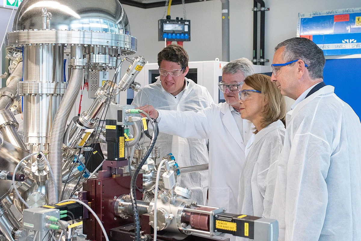 Michael Joukov, Prof. Maximilian Fichtner, Petra Olschowski und Prof. Michael Kühl in weißen Laborkitteln und Laborbrillen in einem HIU-Labor. prof. Fichtner zeigt auf ein Gerät, die Ministerin schaut interessiert