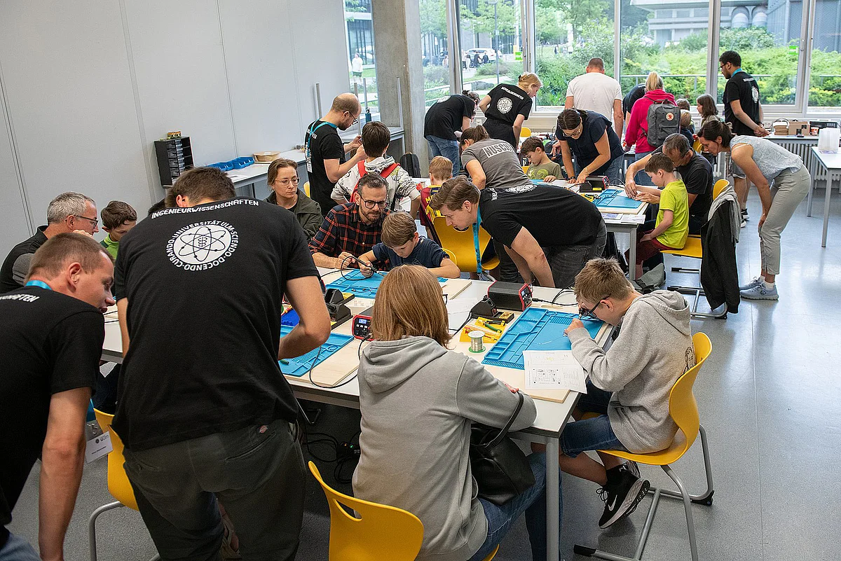 In einem Seminarraum der Uni Ulm sitzen viele Kinder an mehreren Tischen und löten. Dabei werden Sie von Uni-Mirgliedern betreut