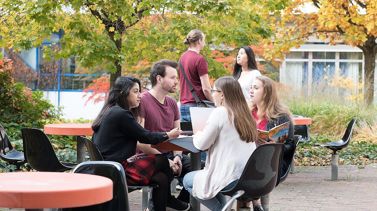 Studierende sitzen in einer Gruppe um einen Tisch im Freien zwei stehen und unterhalten sich