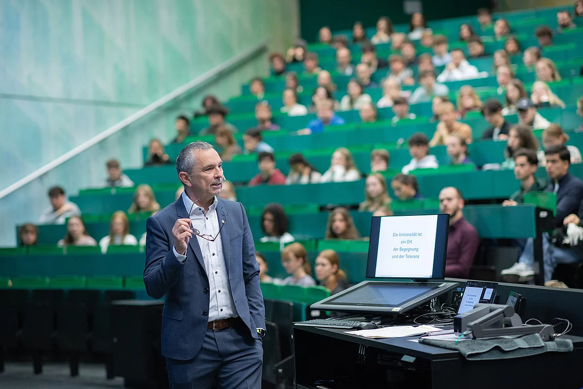In a large lecture hall with green walls and chairs, Prof. Kühl welcomes the students