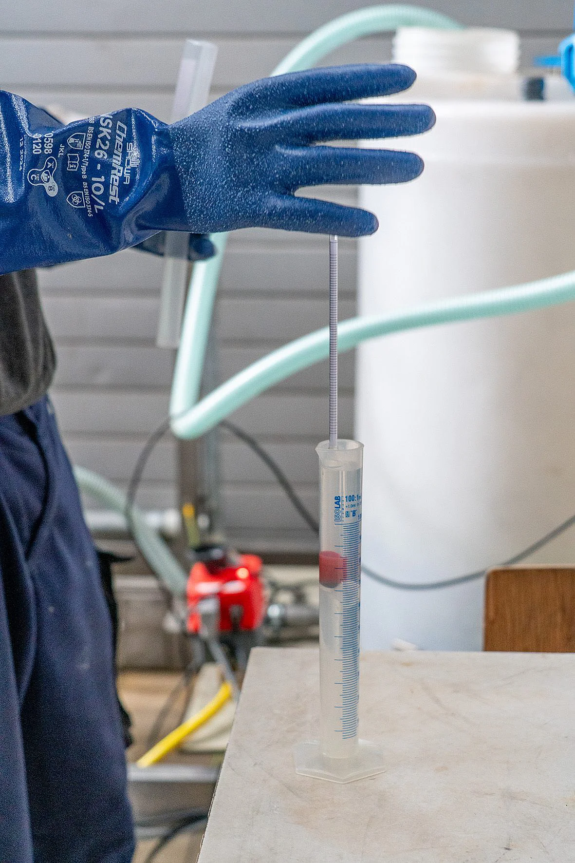 A hand wearing a blue glove drops a plunger into a glass tube containing liquid