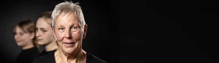 Porträt einer älteren Frau mit kurzen grauen Haaren, zwei jüngere Frauen unscharf im Hintergrund.