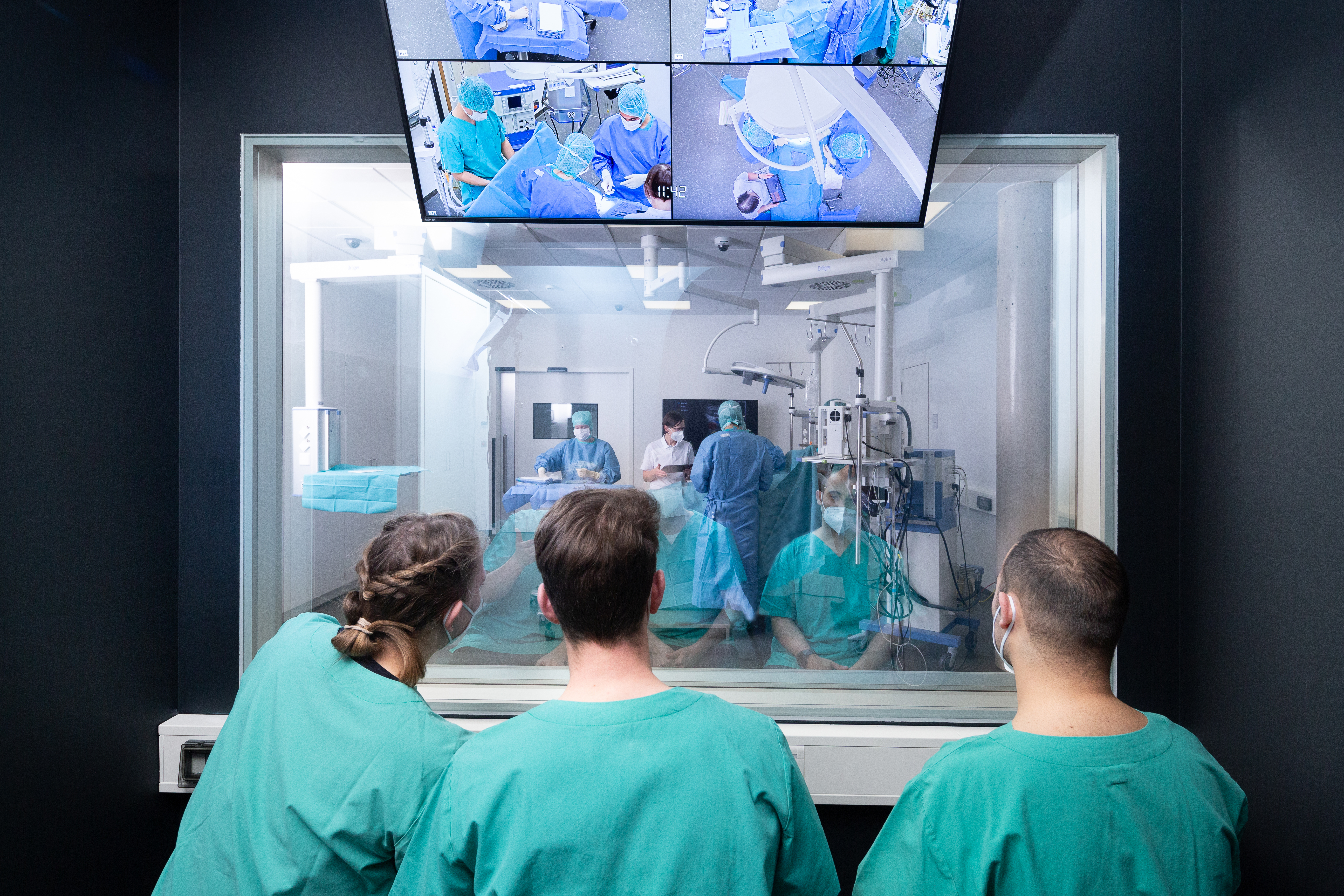 Drei Medizinstudierende sitzen vor einem Operationssaal des Trainingshospitals und schauen einer simulierten Operation zu