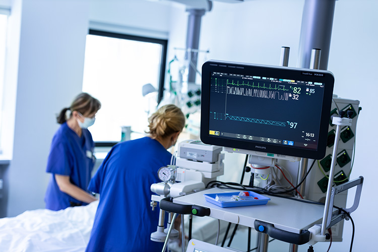  	EKG-Monitor im Vordergrund, medizinisches Fachpersonal betreut einen bettlägerigen Patienten im Hintergrund