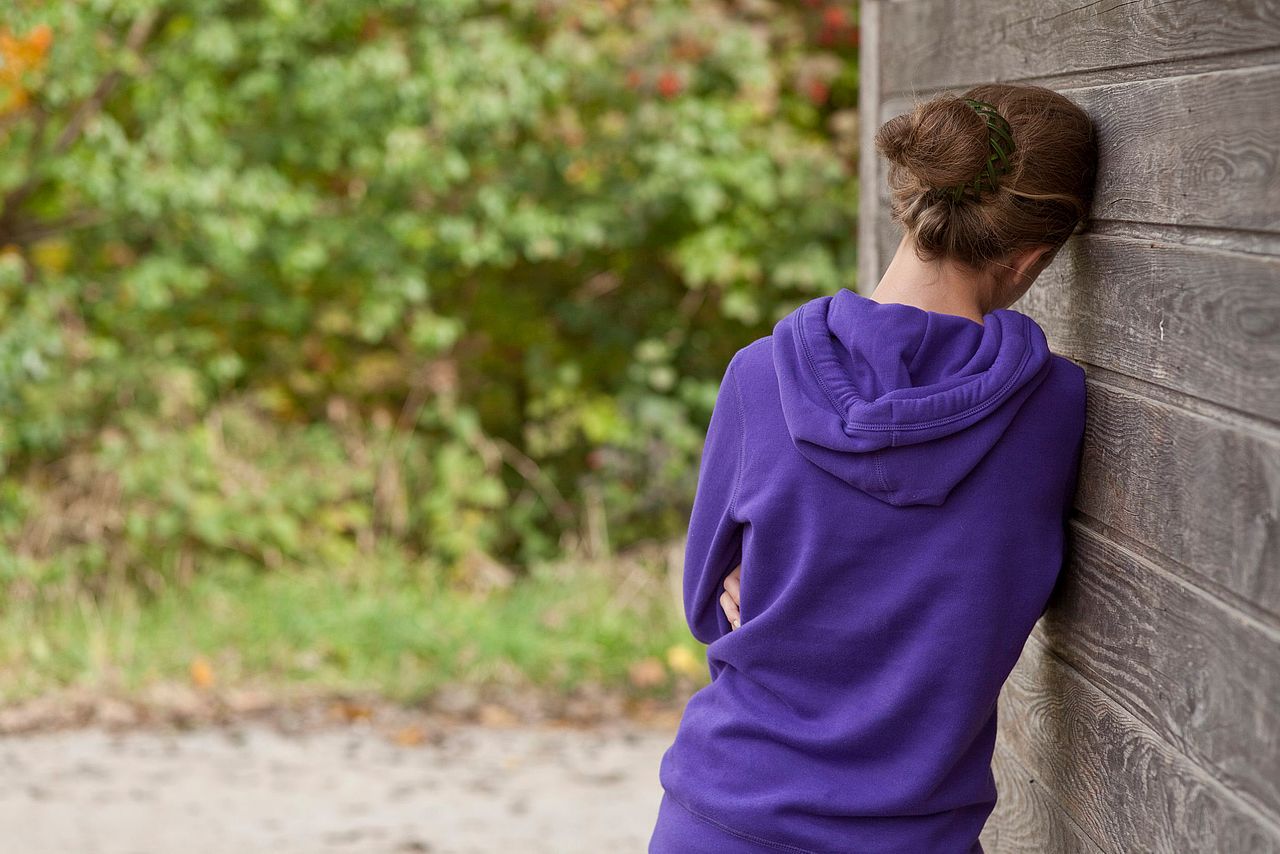 The picture shows a sad looking girl from behind, leaning on a wooden wall.
