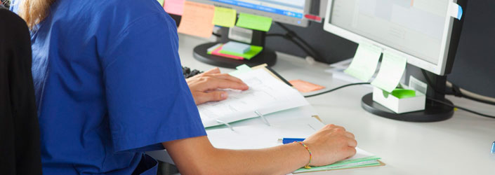 Eine Mitarbeiterin in blauer medizinischer Berufskleidung sitzt am Schreibtisch und bearbeitet Dokumente - Symbolbild für Publikationstätigkeit.