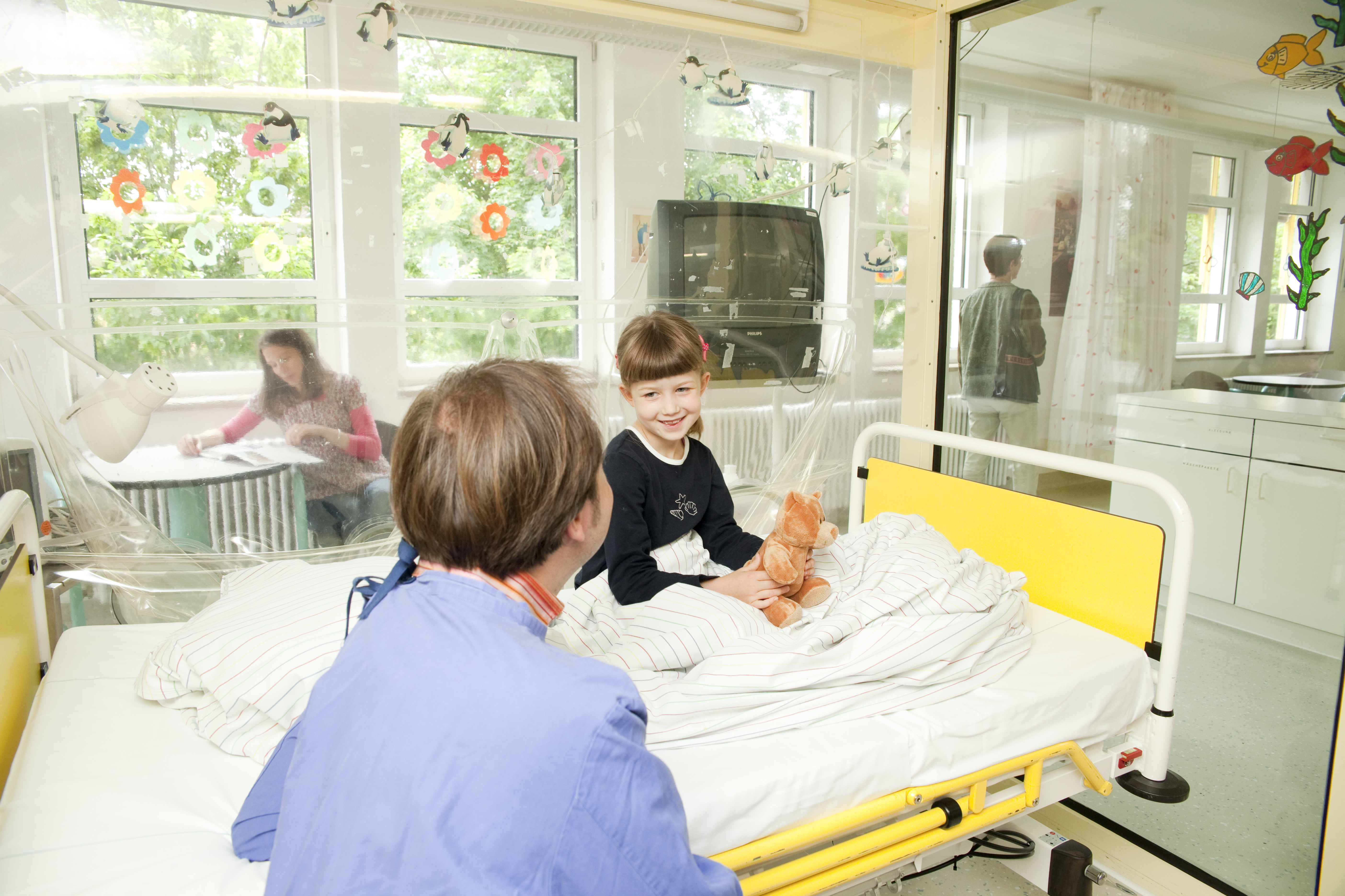 Erwachsener sitzt vor einem Bett auf dem eine junge Patientin sitzt und lacht