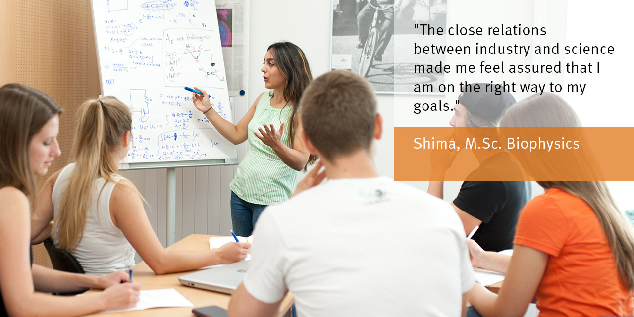 Eine Gruppe Studierender in einem Seminarraum. Eine Studentin präsentiert an einer Flipchart.