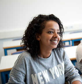 A dark-skinned student in a seminar room. She has medium-length dark, frizzy hair.