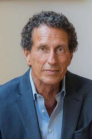 Prof. Dr. Dr. H. C. Julian Nida-Rümelin trägt ein dunkelblaues Sakko über einem hellblauen, leicht geöffneten Hemd. Der Oberkörper ist in einer Nahaufnahme vor einem neutralen, hellen Hintergrund zu sehen. Das Haar ist dunkel und lockig. Keine weiteren Accessoires oder auffälligen Gegenstände sind sichtbar.