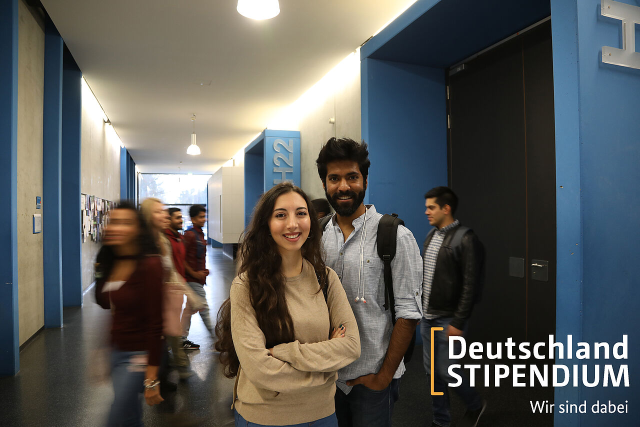 Zwei Studierende stehen vor einem Hörsaal und blicken in die Kamera, während im Hintergrund weitere Studierende vorbei gehen. Das Bild ist auf eine Informationsseite für Bewerber verlinkt.
